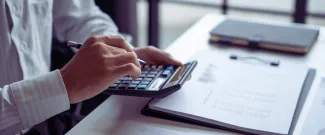 A person working at a desk, holding a pen while typing on a calculator, with a clipboard and notebook nearby.