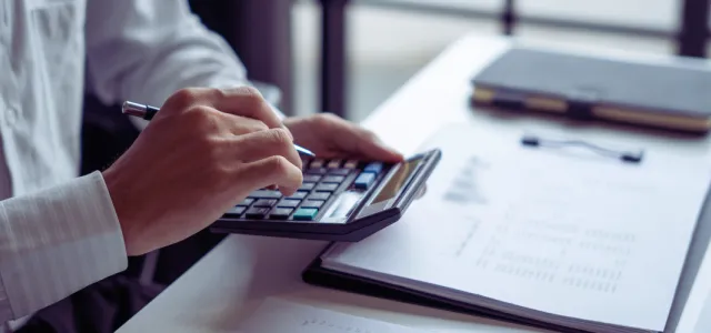 A person working at a desk, holding a pen while typing on a calculator, with a clipboard and notebook nearby.